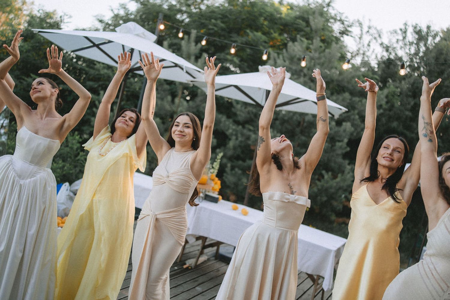 Wedding party wearing yellow and beige dresses pose joyfully with raised arms in an outdoor garden setting.
