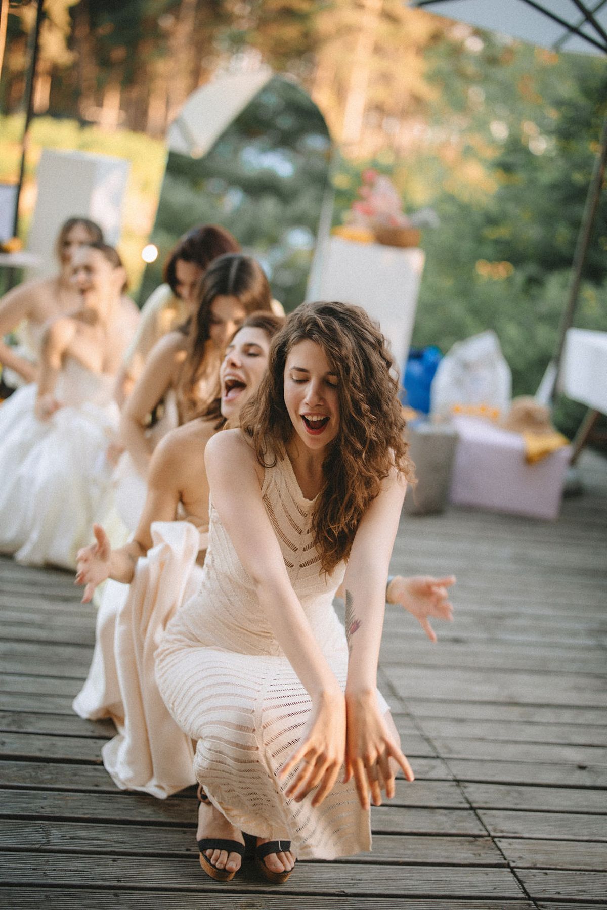 A group in light-colored dresses laughs joyfully while sitting on a wooden deck during a summer evening.