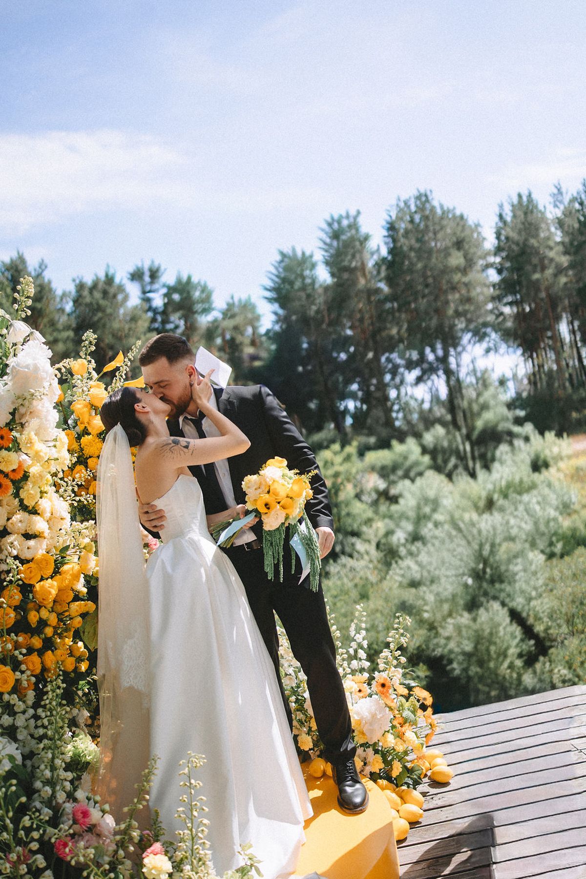 Romantic outdoor wedding ceremony with bright yellow sunflower decorations on a wooden deck surrounded by lush greenery.
