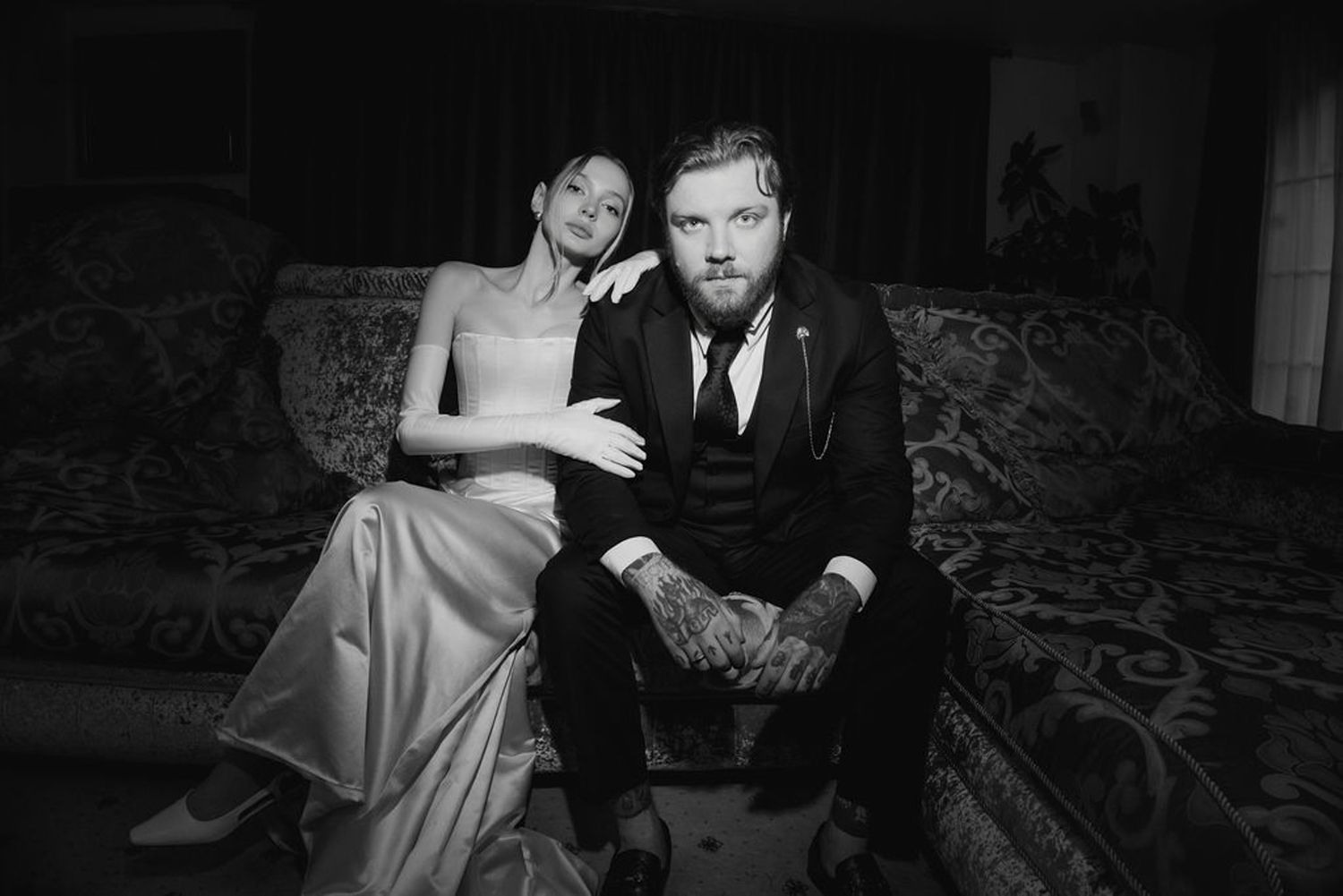 A couple sits together on an ornate couch in a black and white photograph.