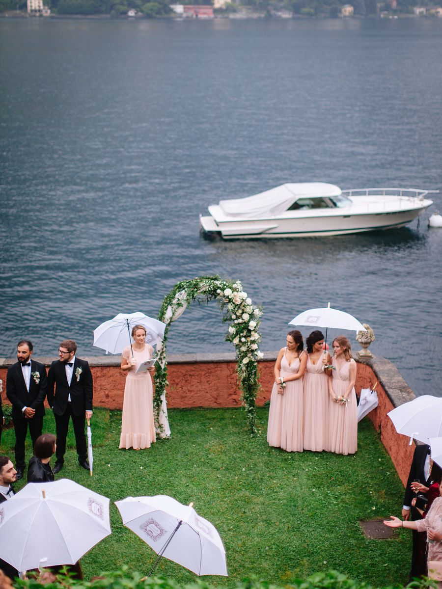 A wedding party with umbrellas gathers on a grassy lawn overlooking a serene lake on an overcast day.