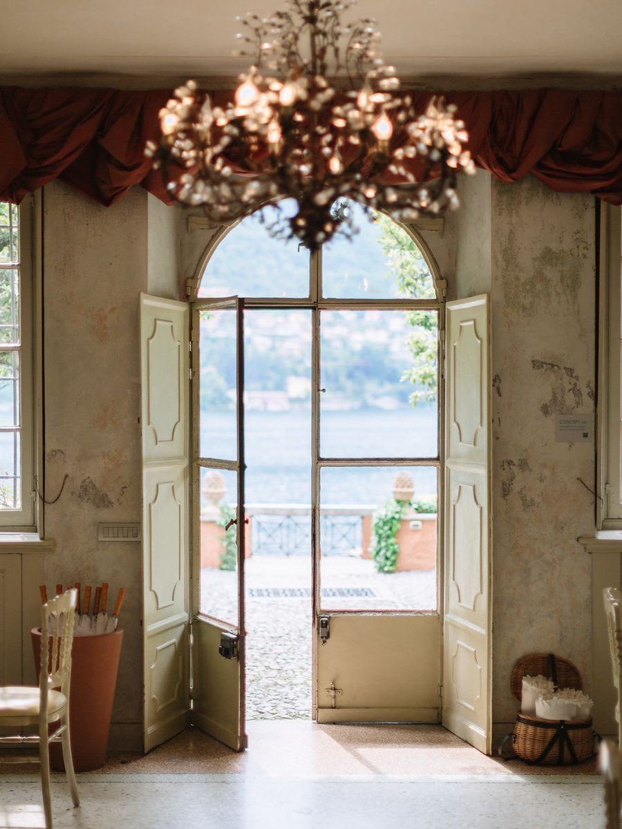 Elegant French doors with arched windows open to bright daylight under an ornate chandelier in a vintage interior.