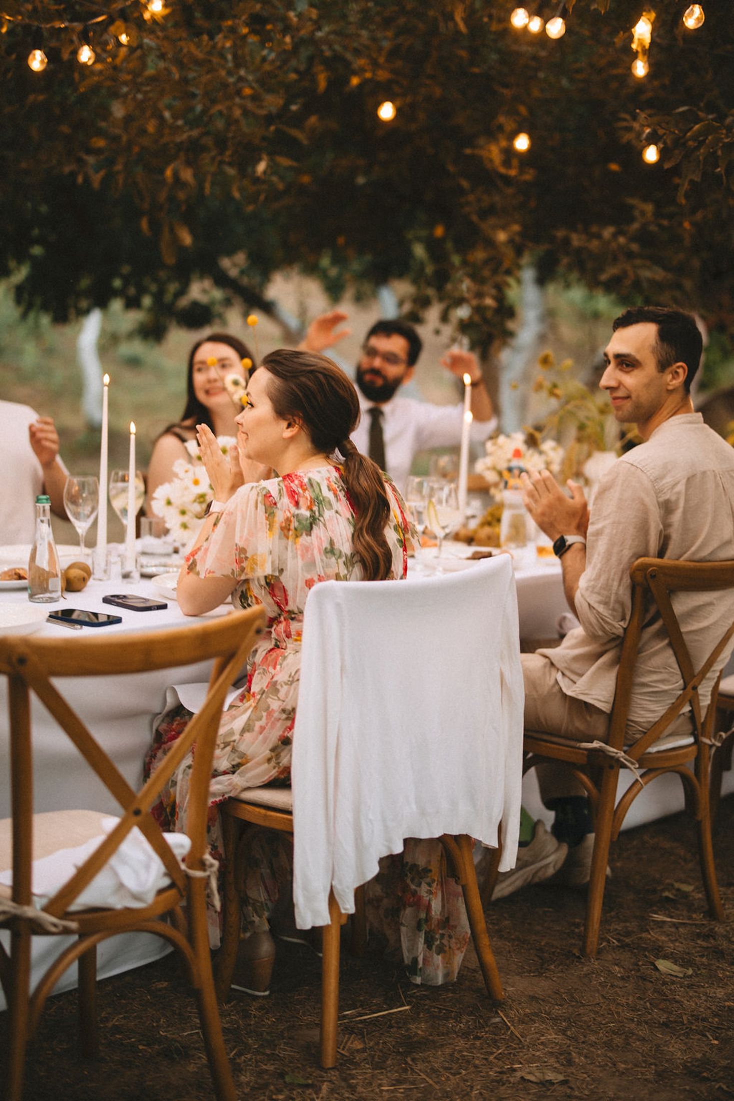 Guests enjoy an intimate outdoor dinner party under string lights and trees at dusk with rustic wooden chairs and white linens.