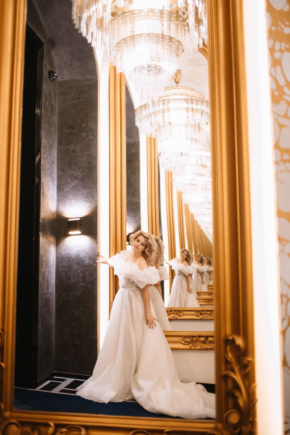 Bride in flowing white gown poses elegantly beneath crystal chandeliers in ornate hallway.
