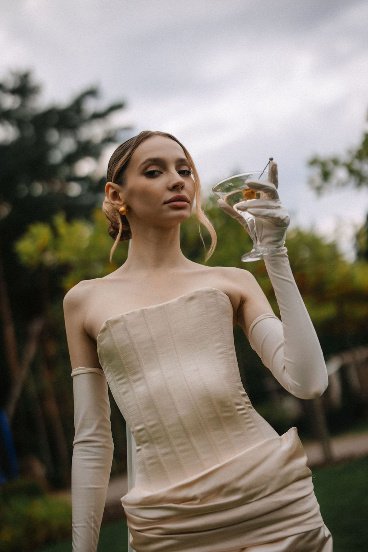 Artistic fashion portrait showing a model in a cream-colored corset top with long sleeves against a cloudy outdoor backdrop.