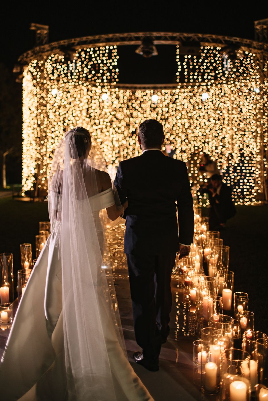 A couple walks down a candlelit aisle with a stunning wall of twinkling lights behind them.