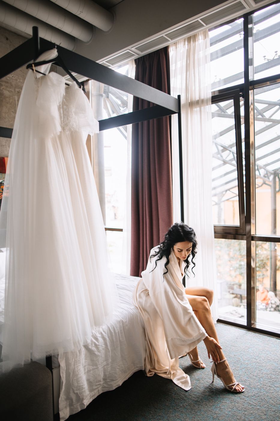 A bridal gown hangs elegantly in a modern room with floor-to-ceiling windows and soft natural lighting.