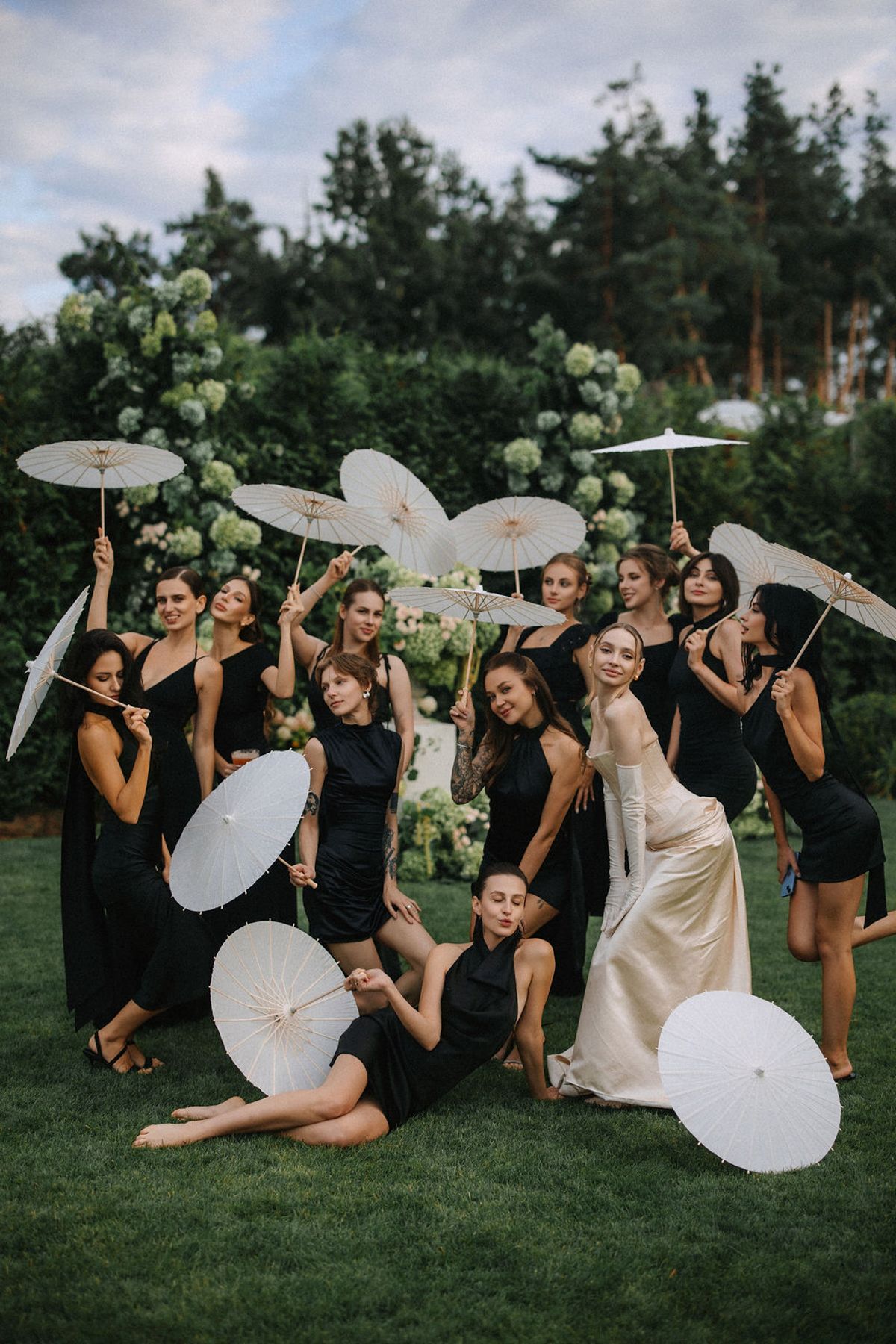 A wedding party in black dresses poses playfully with white paper parasols on a green lawn at dusk.