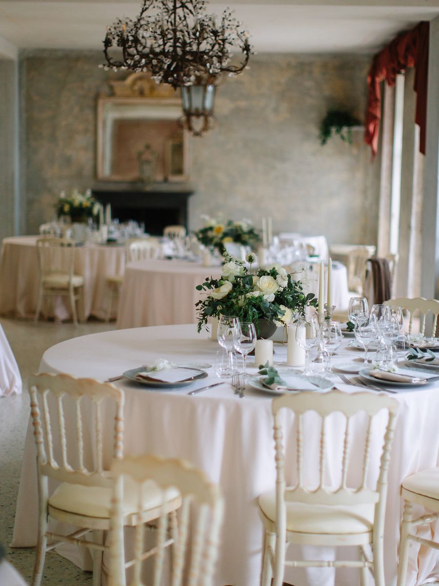 Elegant wedding reception table setting with white linens, floral centerpieces and crystal chandelier in stone-walled room.