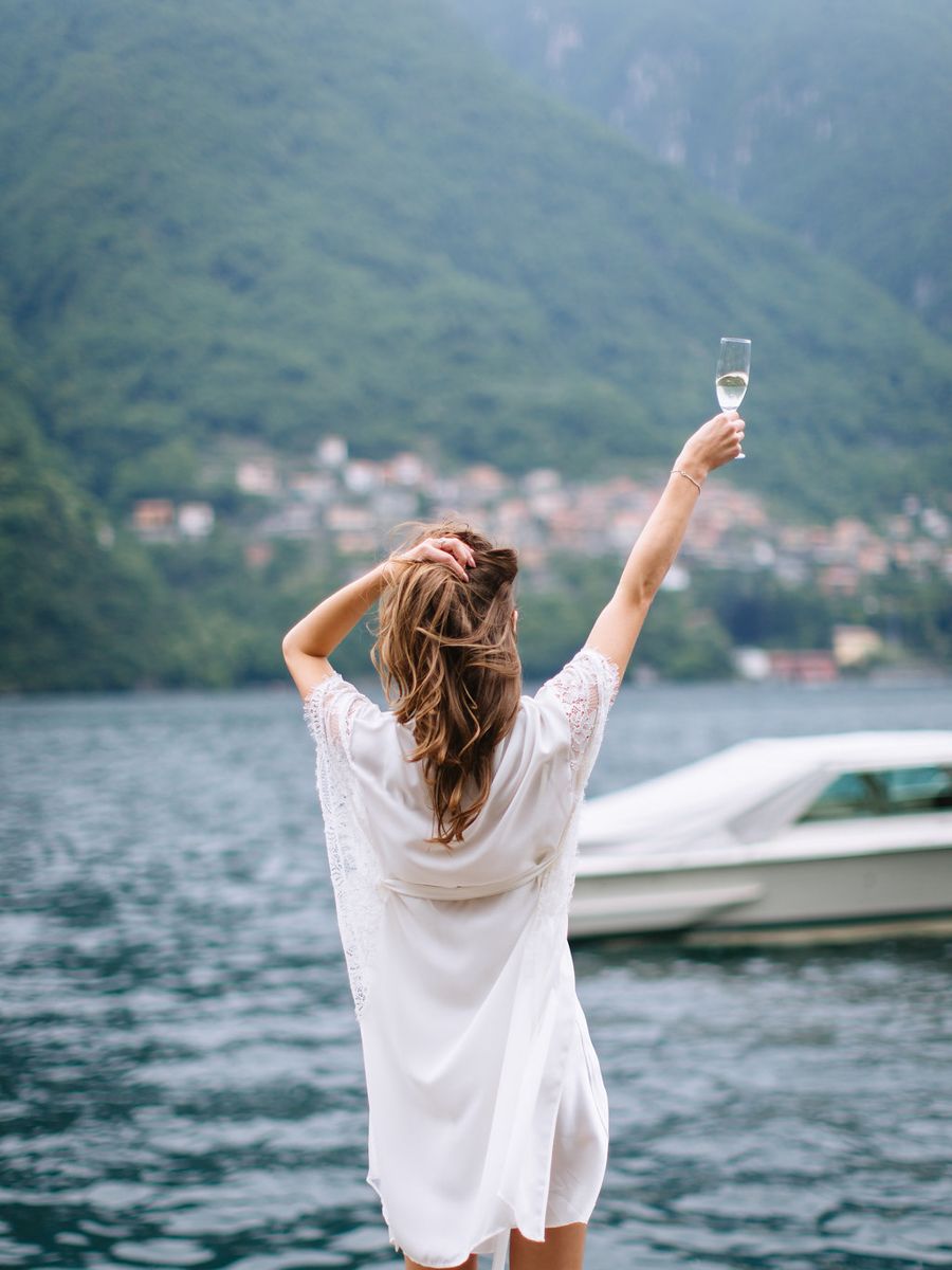 Woman in white dress overlooking scenic mountain lake with boats and village in background on sunny day.
