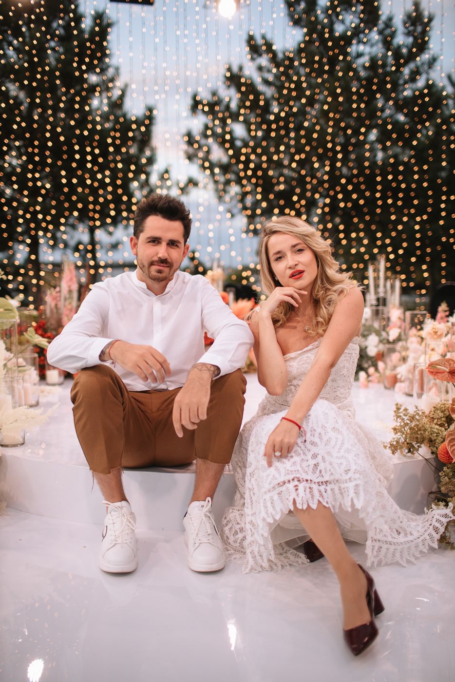 A couple sits on a white surface surrounded by twinkling fairy lights and floral decorations at an elegant event.