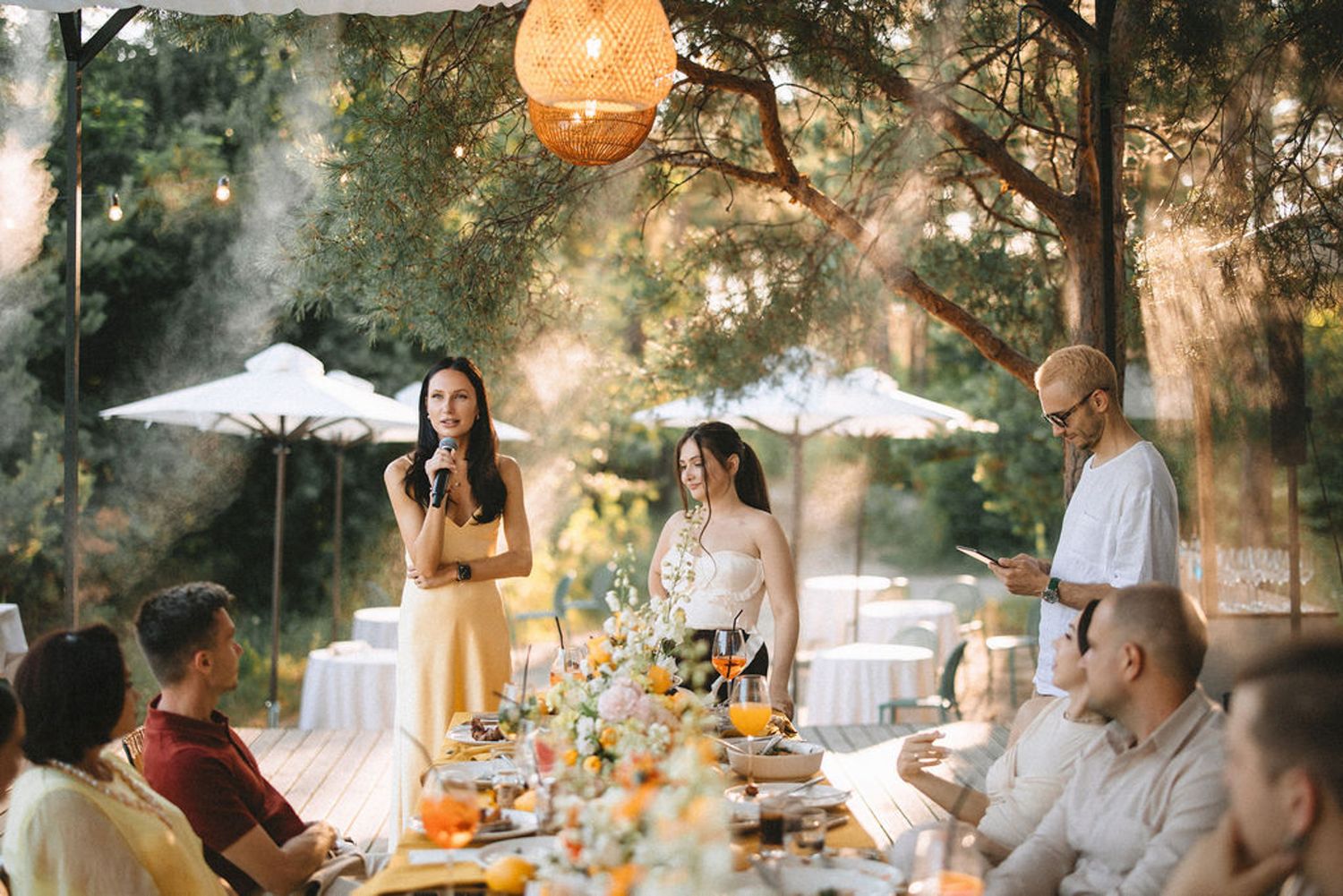 A festive outdoor dinner party setup with string lights, umbrellas and tables arranged under trees at dusk.
