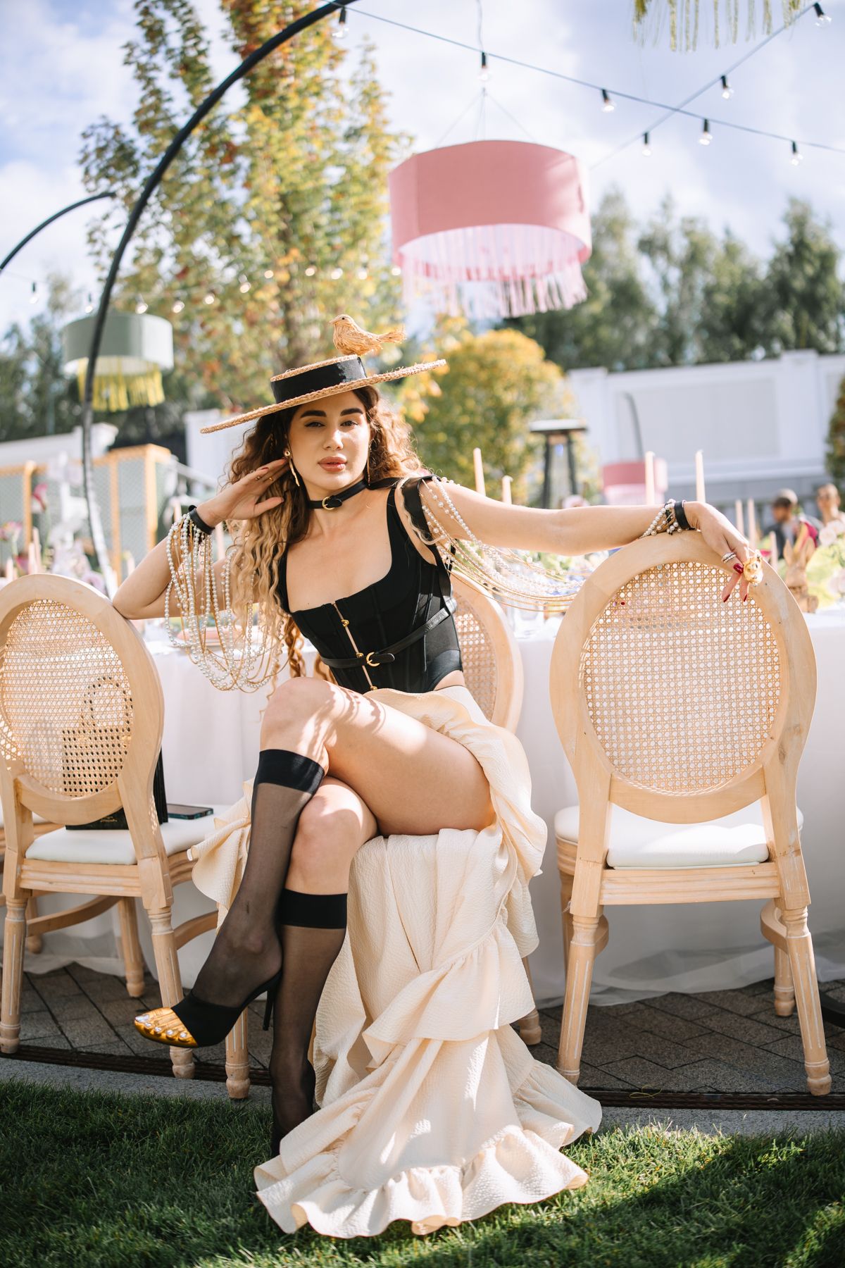 A figure in a cream dress and sun hat poses gracefully across vintage cane chairs in an outdoor garden setting.