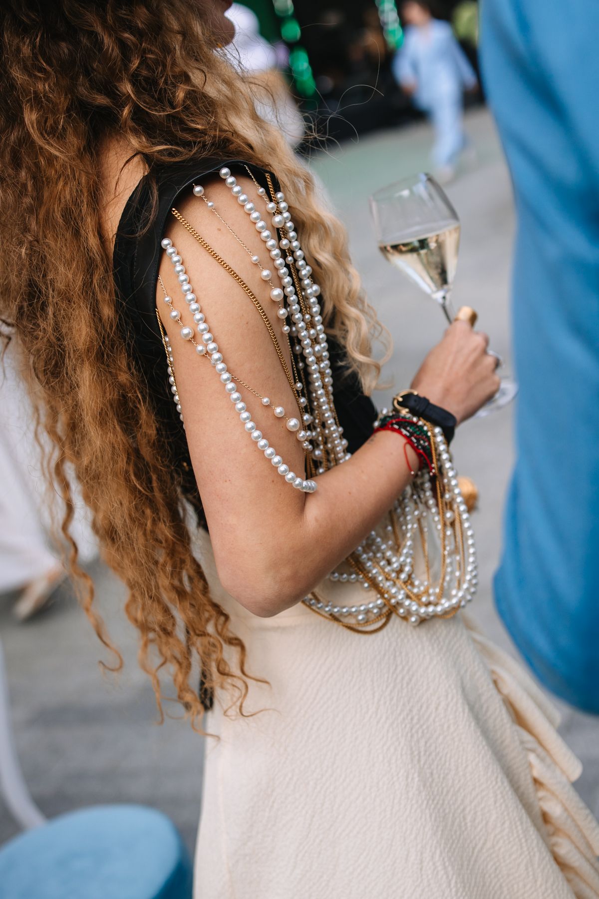 Person with curly hair wearing a white dress and layered pearl necklaces at an outdoor event.