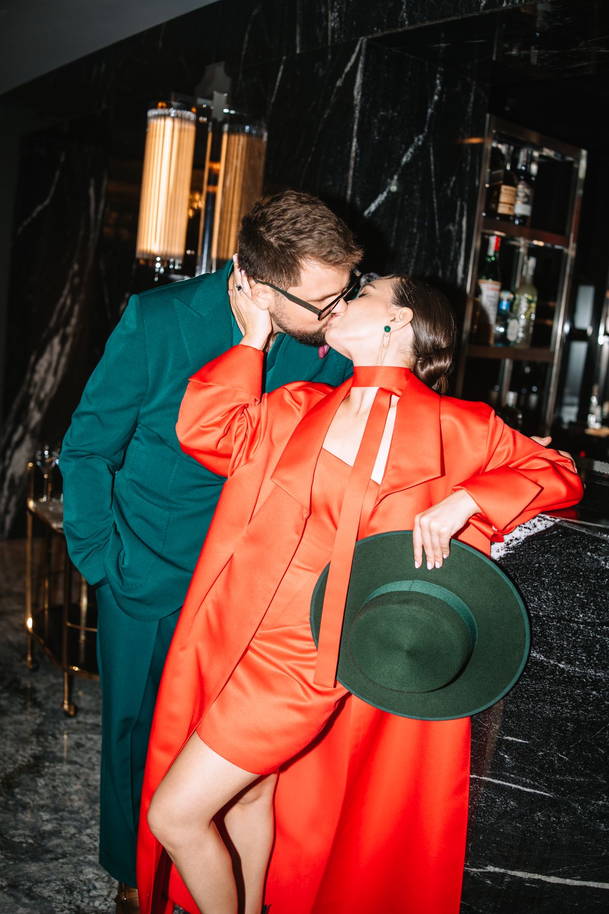 Two people in contrasting orange and green outfits embrace in a dimly lit room with black marble walls.