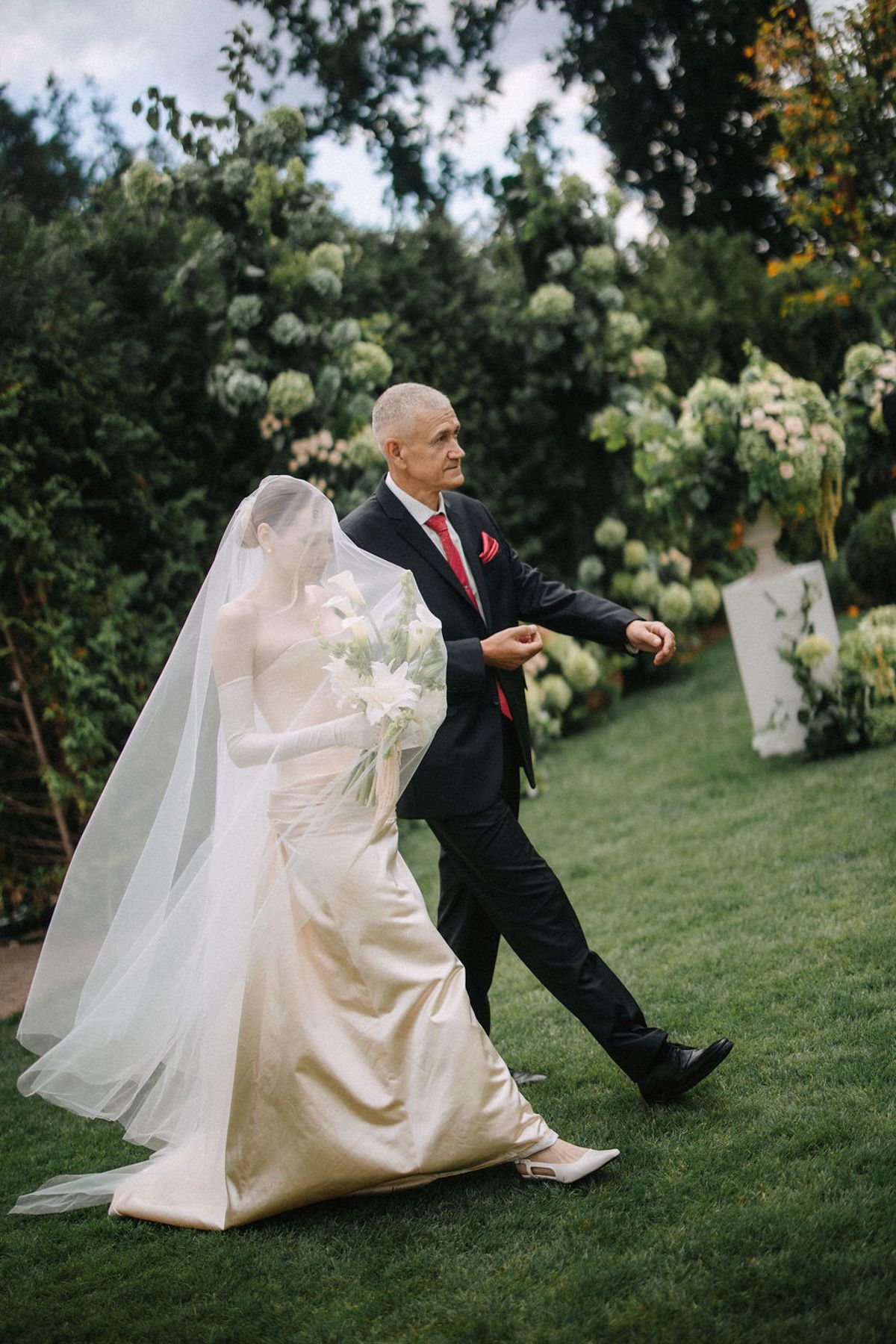 A bride in a flowing white dress and veil dances playfully with someone in a dark suit on a grassy lawn with flowers.