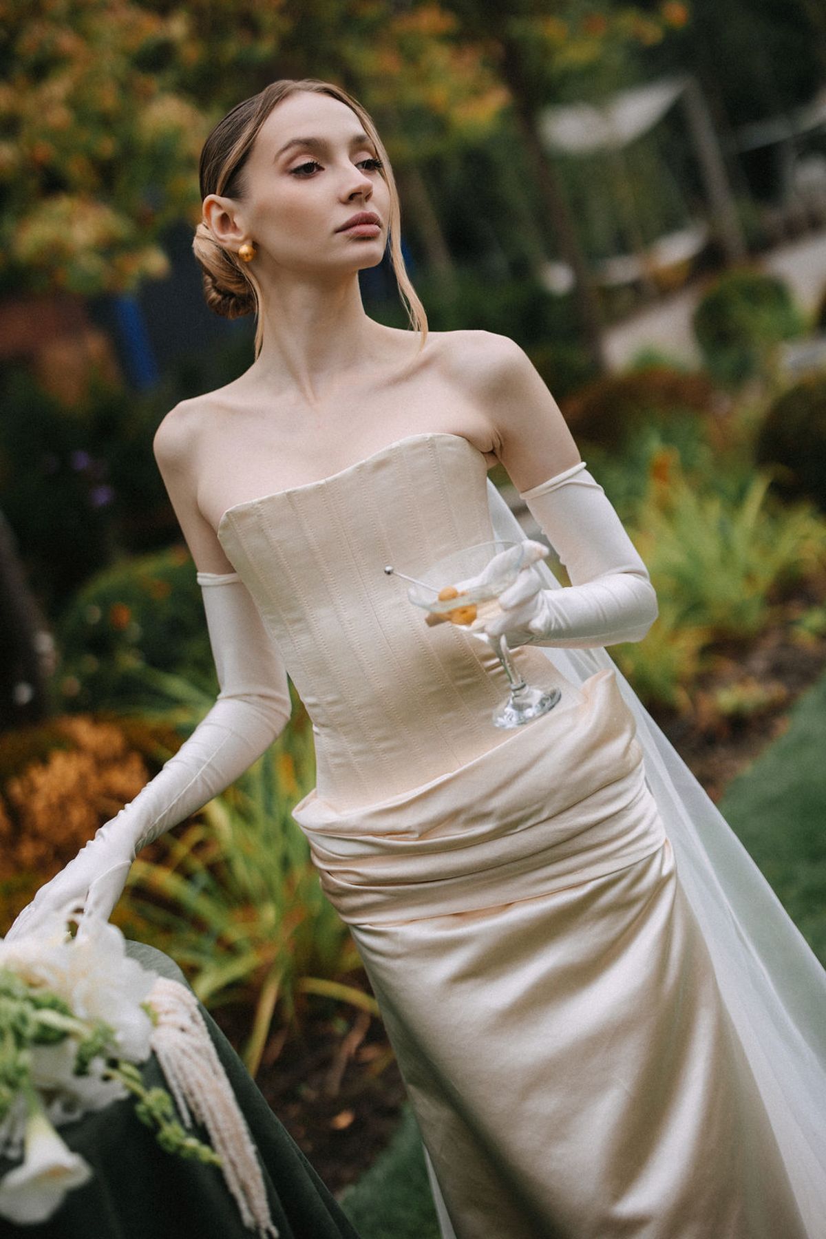 A bride in an elegant off-shoulder cream dress with long gloves poses gracefully in a garden setting.