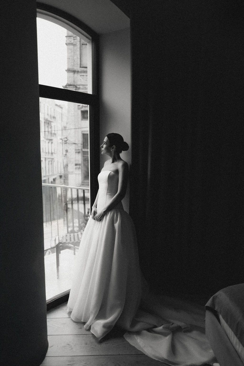 Black and white photo of a bride in a flowing wedding gown gazing out large windows in an elegant interior.