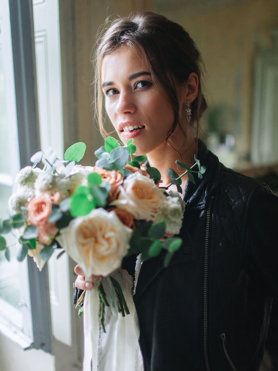 A stunning bridal bouquet of peach garden roses and greenery is held near a window in soft natural light.