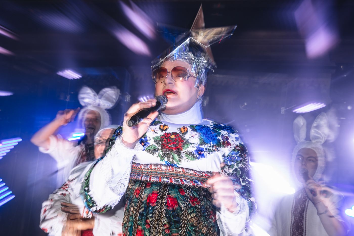 Performer wearing traditional embroidered folk costume dances under purple stage lights.