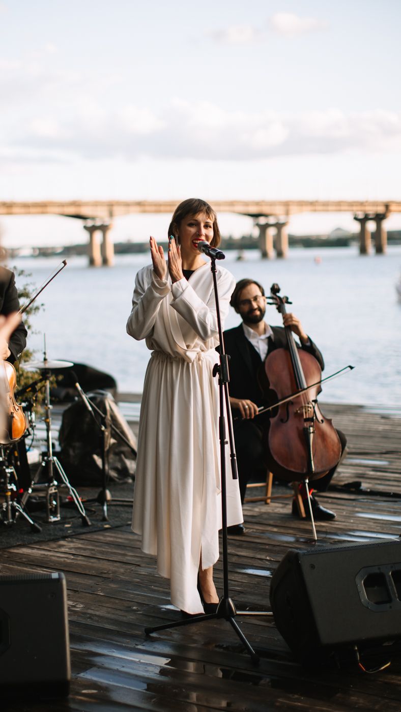 Musicians perform at a waterfront venue with a cellist and vocalist on a wooden dock at sunset.