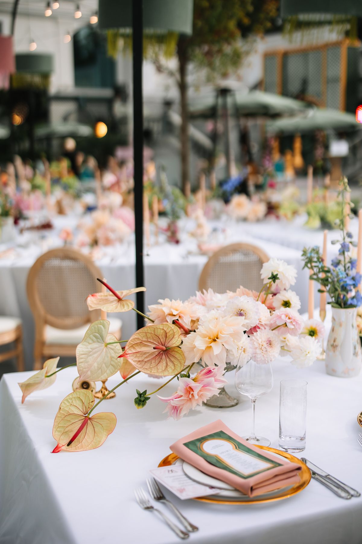 Elegant wedding reception table with pink floral centerpieces, anthurium leaves, and vintage French chairs at an outdoor venue.