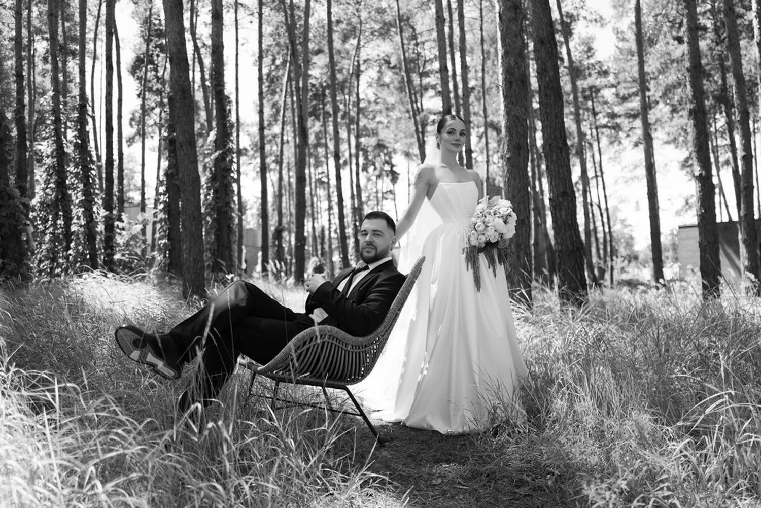 Black and white wedding photo series showing couple in formal attire posing romantically among tall pine trees in forest.