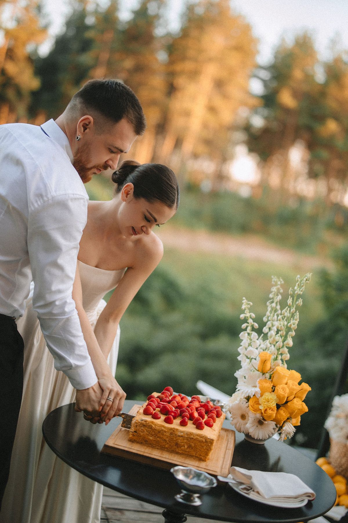 A couple in wedding attire cutting into a rustic wedding cake decorated with fresh berries and flowers at sunset.