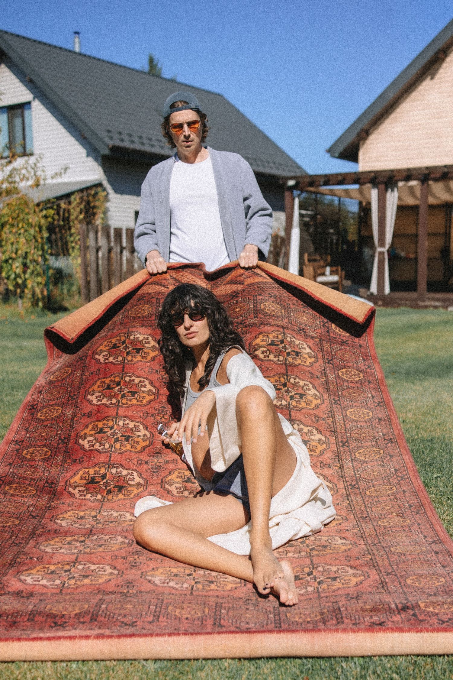 A couple lounges together on a decorative Persian rug laid out on grass in a backyard setting with houses visible.