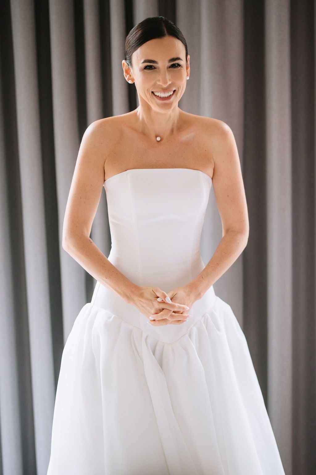 A person wearing a strapless white wedding gown smiles warmly while standing against a gray curtain background.