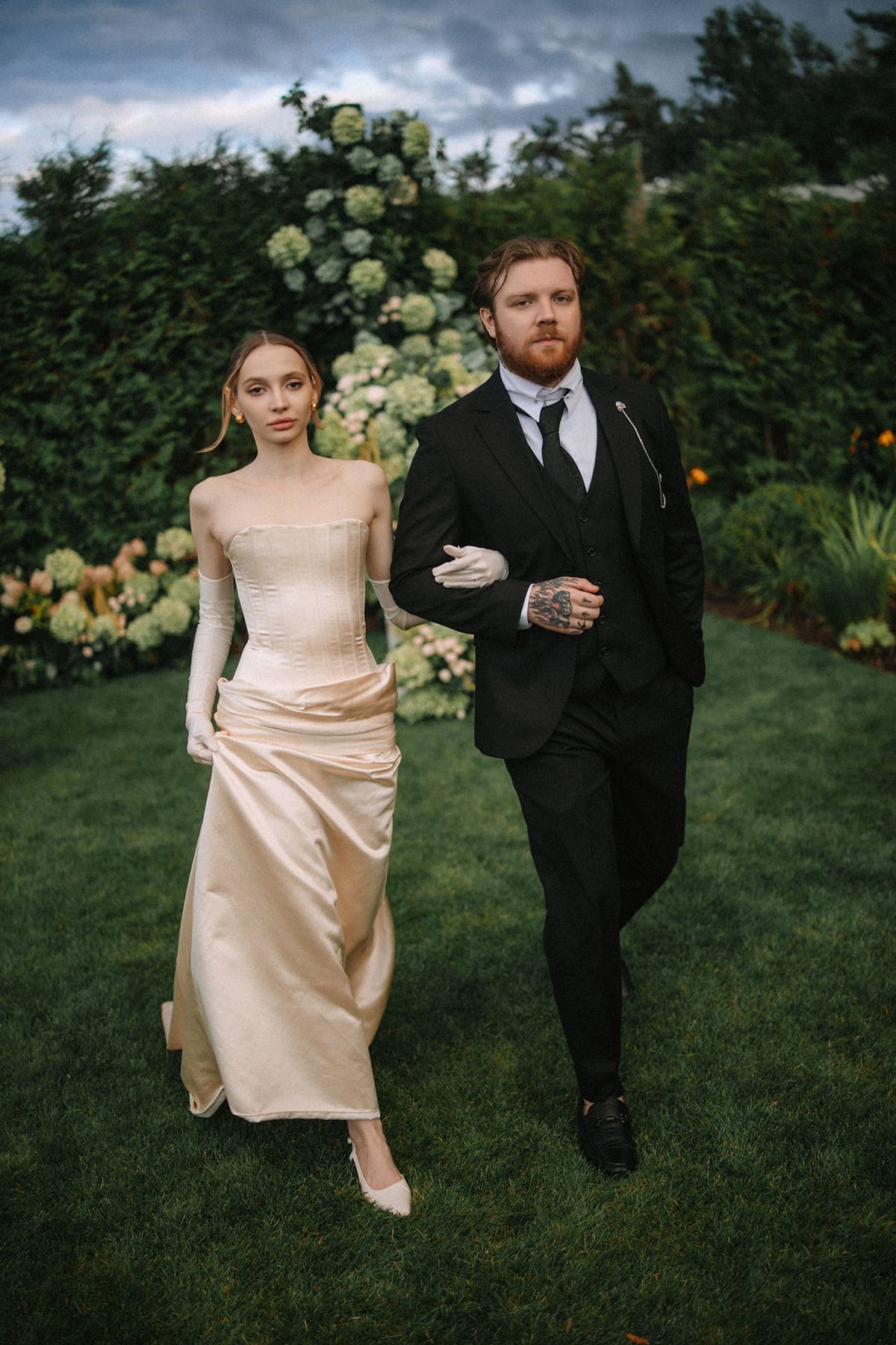 A couple in wedding attire walks together on a lush green lawn during an outdoor celebration.