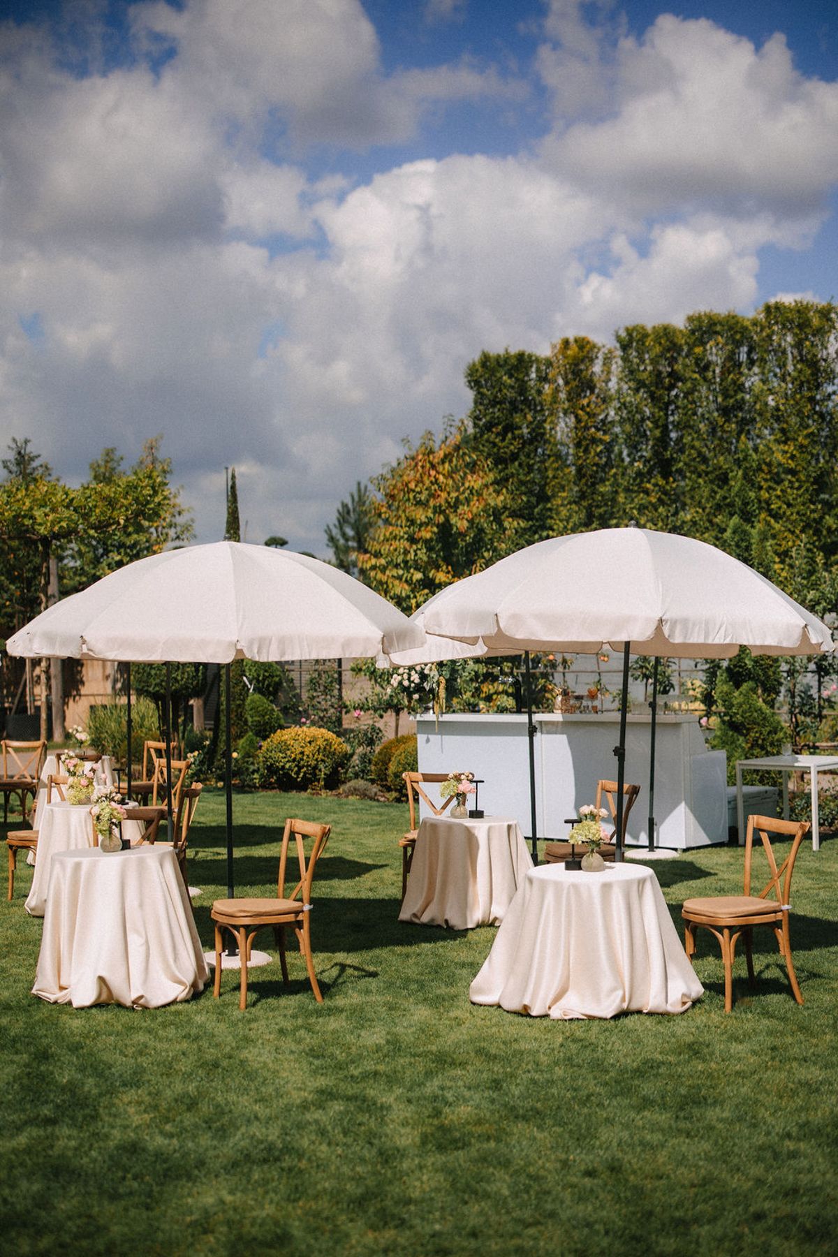 Elegant outdoor event setup with white umbrellas, round tables with tablecloths, and wooden chairs on a manicured lawn.