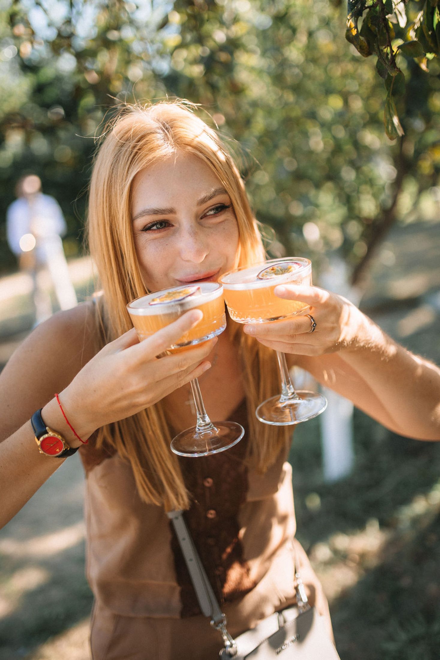 Person in brown dress toasting with cocktail glasses outdoors in a sunny garden setting.