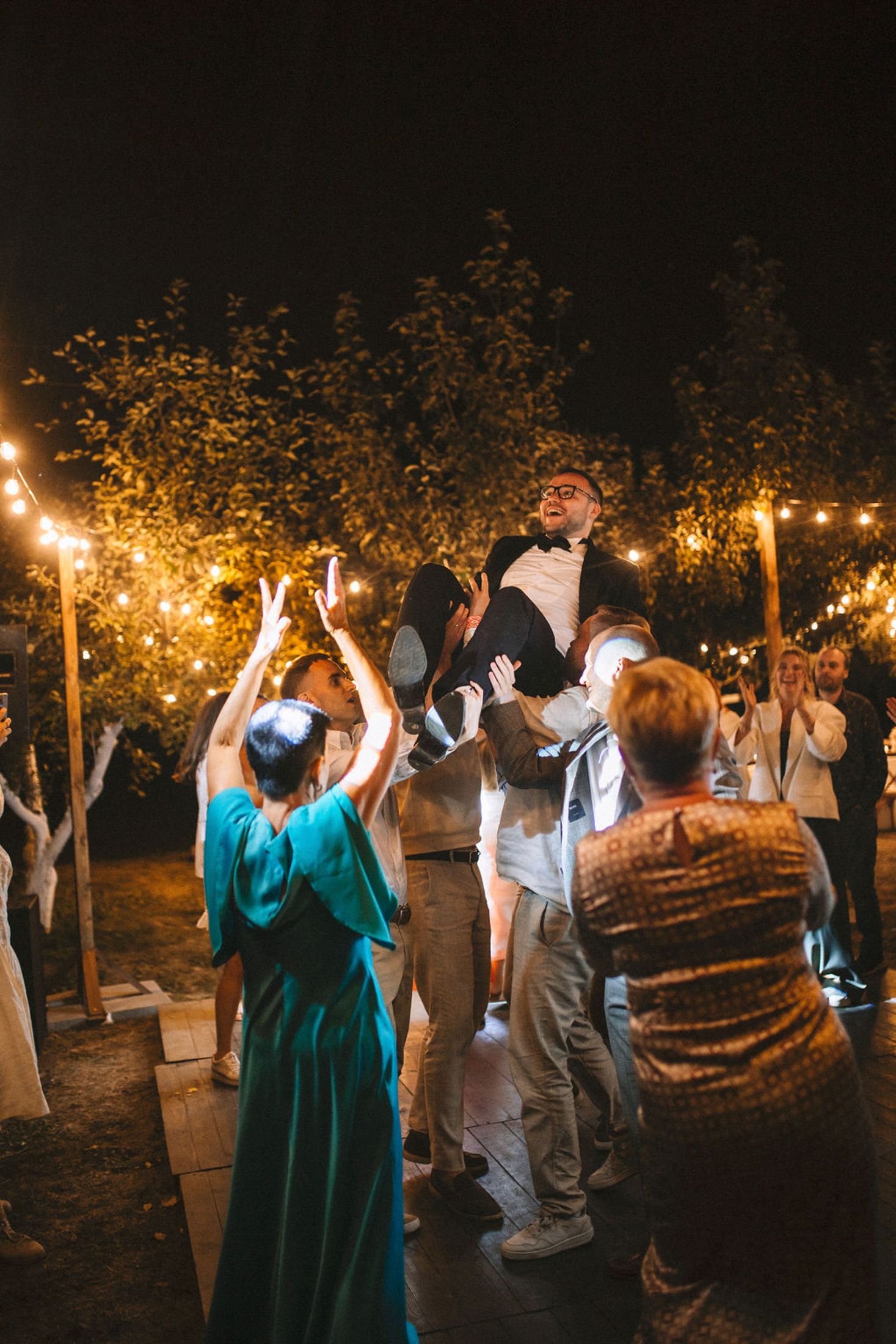 Guests celebrate at an outdoor nighttime wedding reception under string lights and trees with someone being lifted in the air.