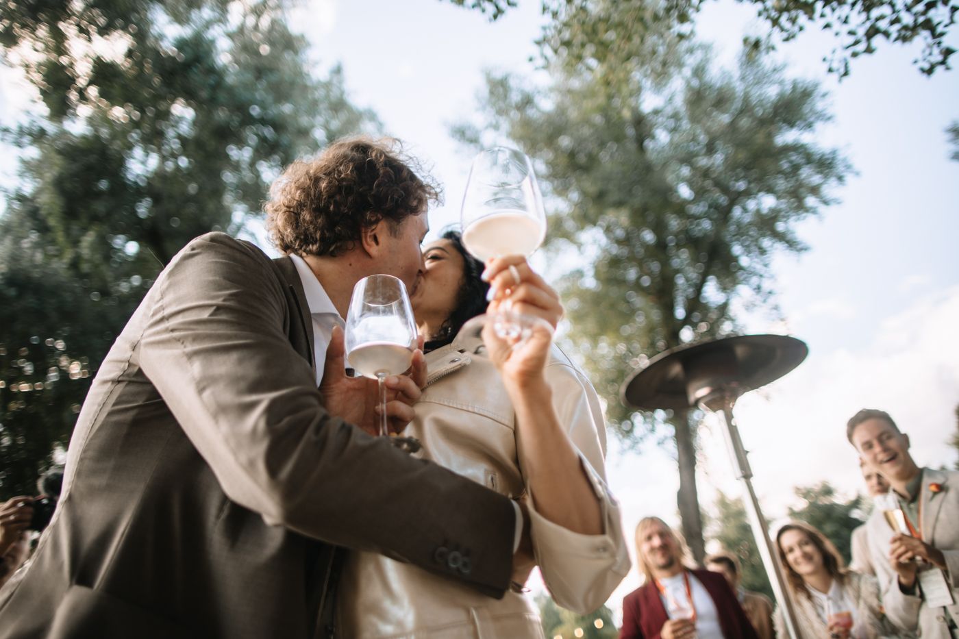 Wedding guests raise their glasses in a celebratory toast during an outdoor reception under trees.
