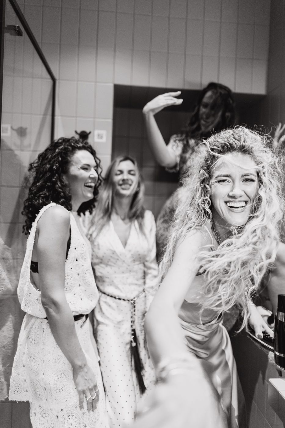A black and white photograph showing a group of women laughing and dancing together in white dresses.