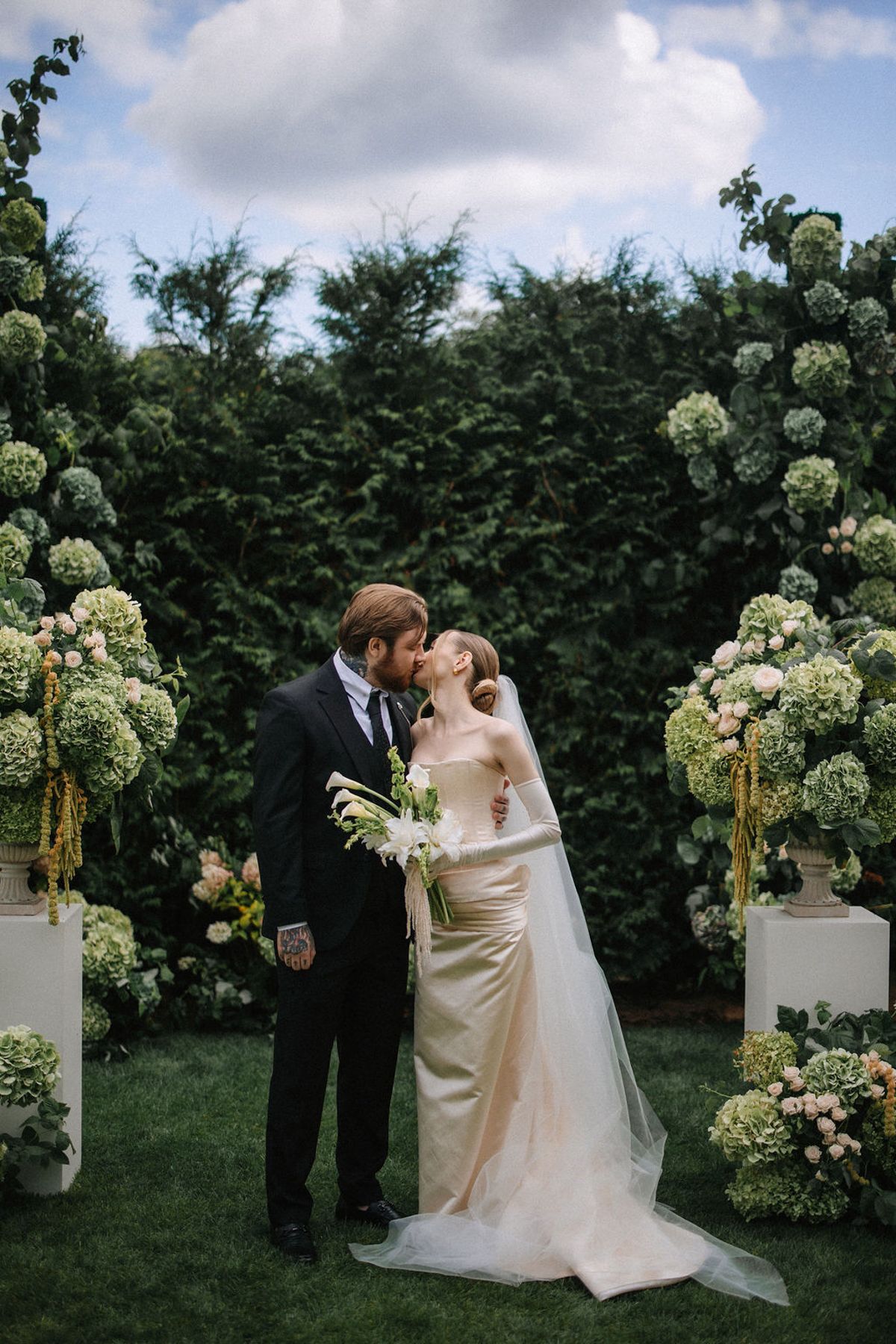 A couple shares an intimate moment at their garden wedding ceremony surrounded by white floral arrangements and greenery.