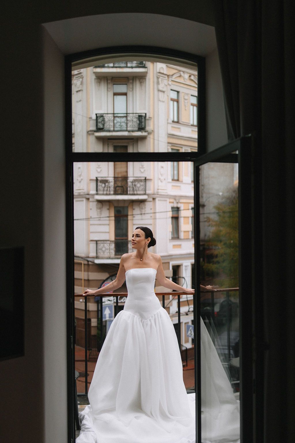 A bride in a strapless white gown stands on a balcony against a backdrop of historic European architecture.