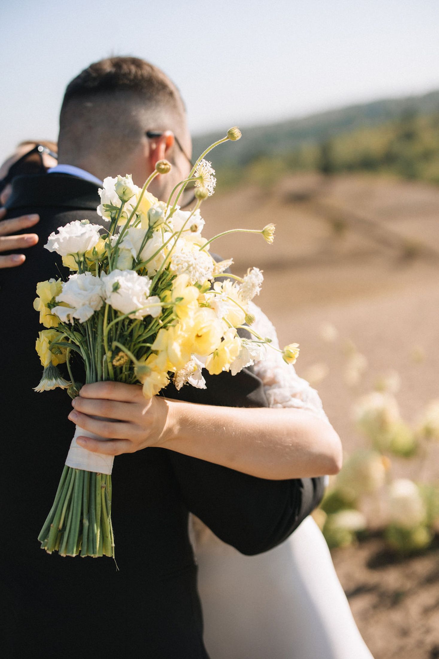 A romantic embrace between newlyweds holding a delicate bouquet of white and yellow flowers in an outdoor setting.