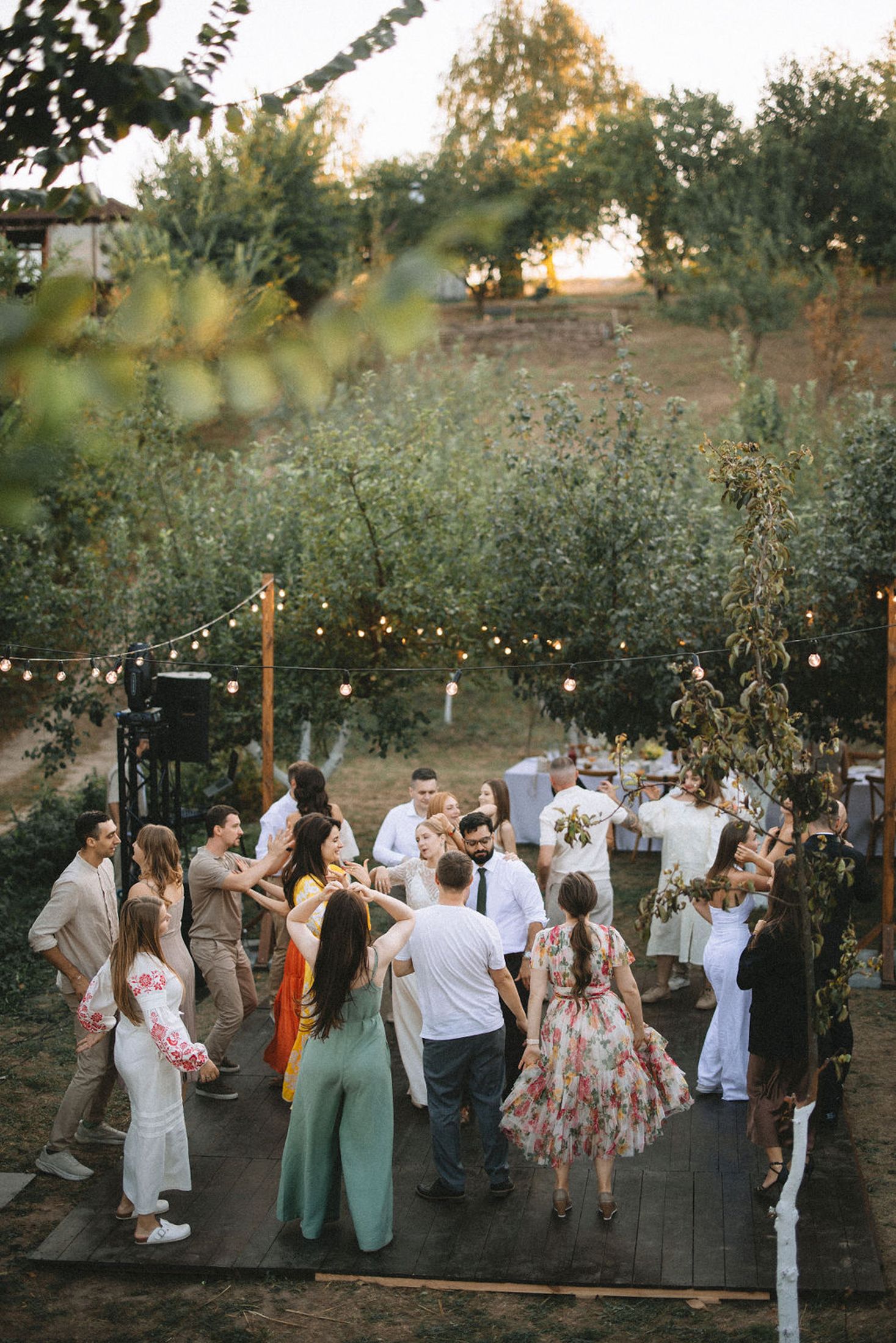 Wedding guests dance together under string lights at an outdoor evening reception in a garden setting.
