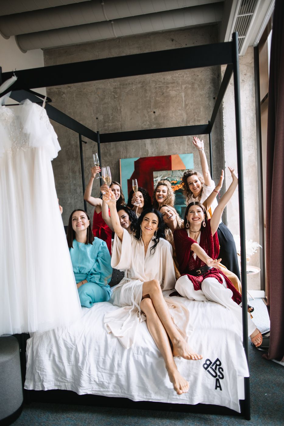 A group of friends in white robes lounge and celebrate together on a bed in a bright room.