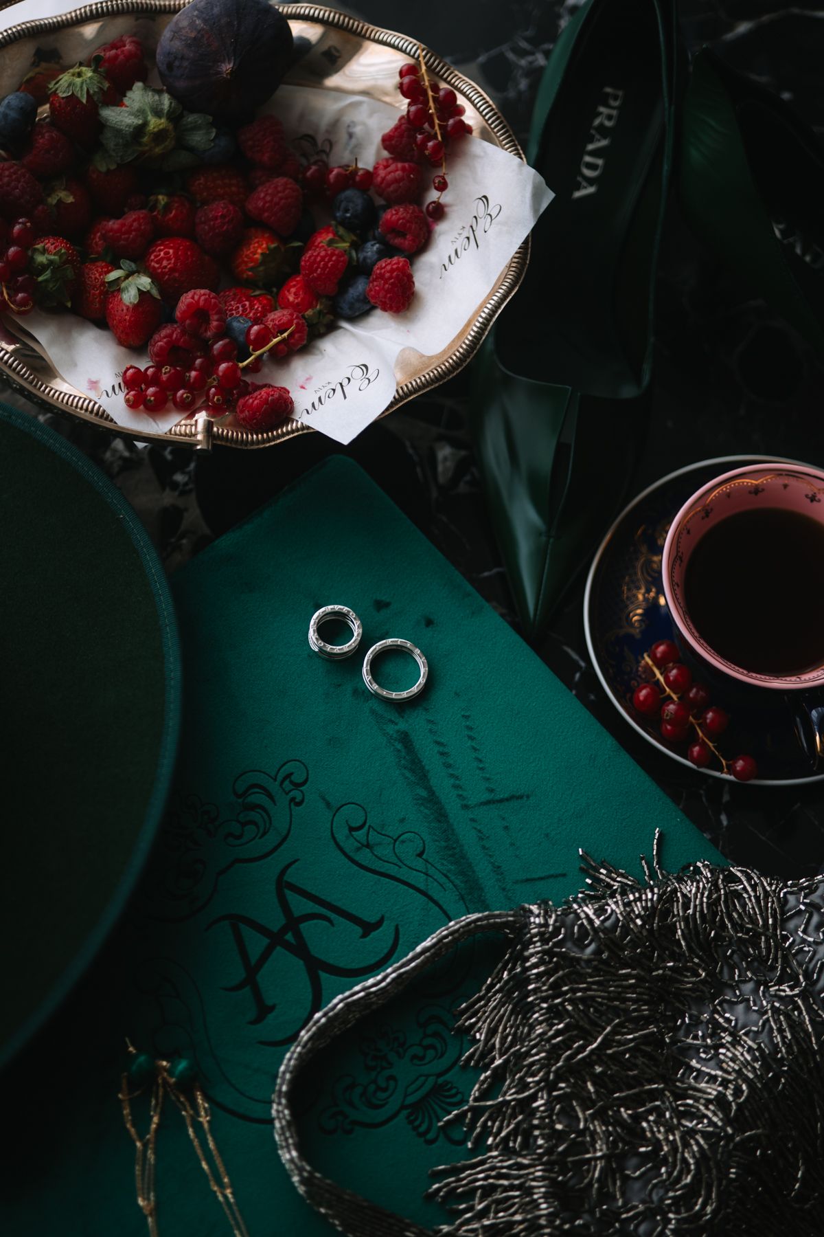 Dark moody still life with fresh red berries and wedding rings on a teal green surface.