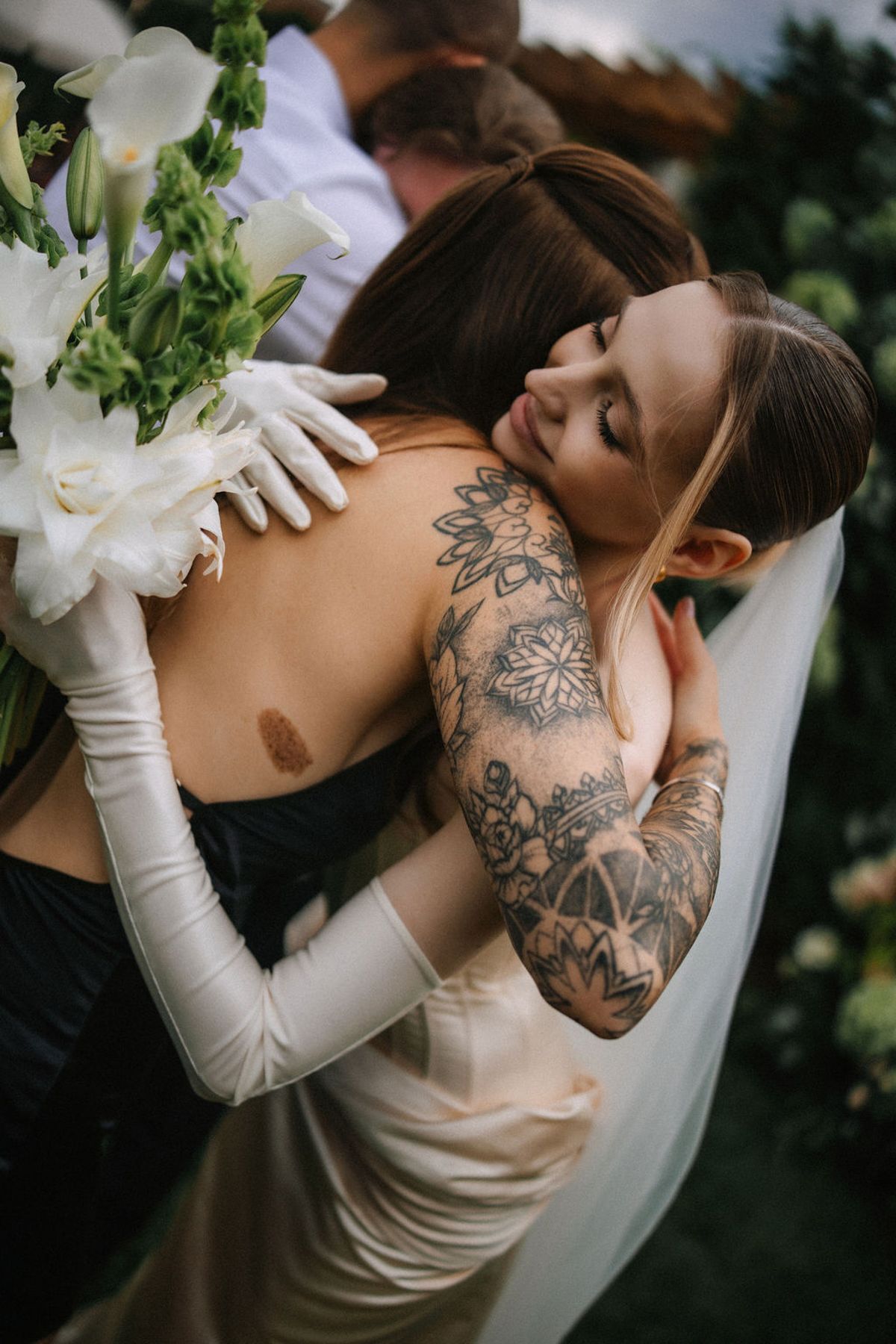 A bride in long white gloves and floral veil embraces her partner while showing intricate floral arm tattoos.