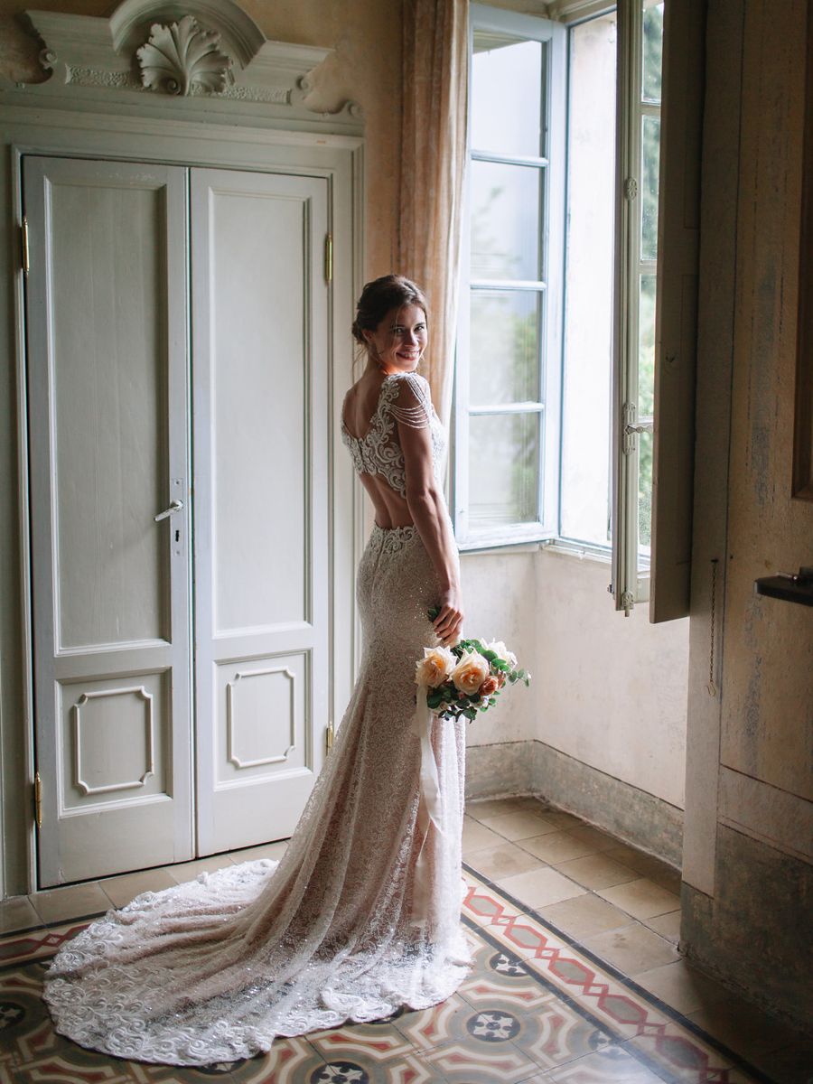 A bride in a vintage lace wedding gown stands by sunlit windows in an elegant room with ornate molding and tiled floors.