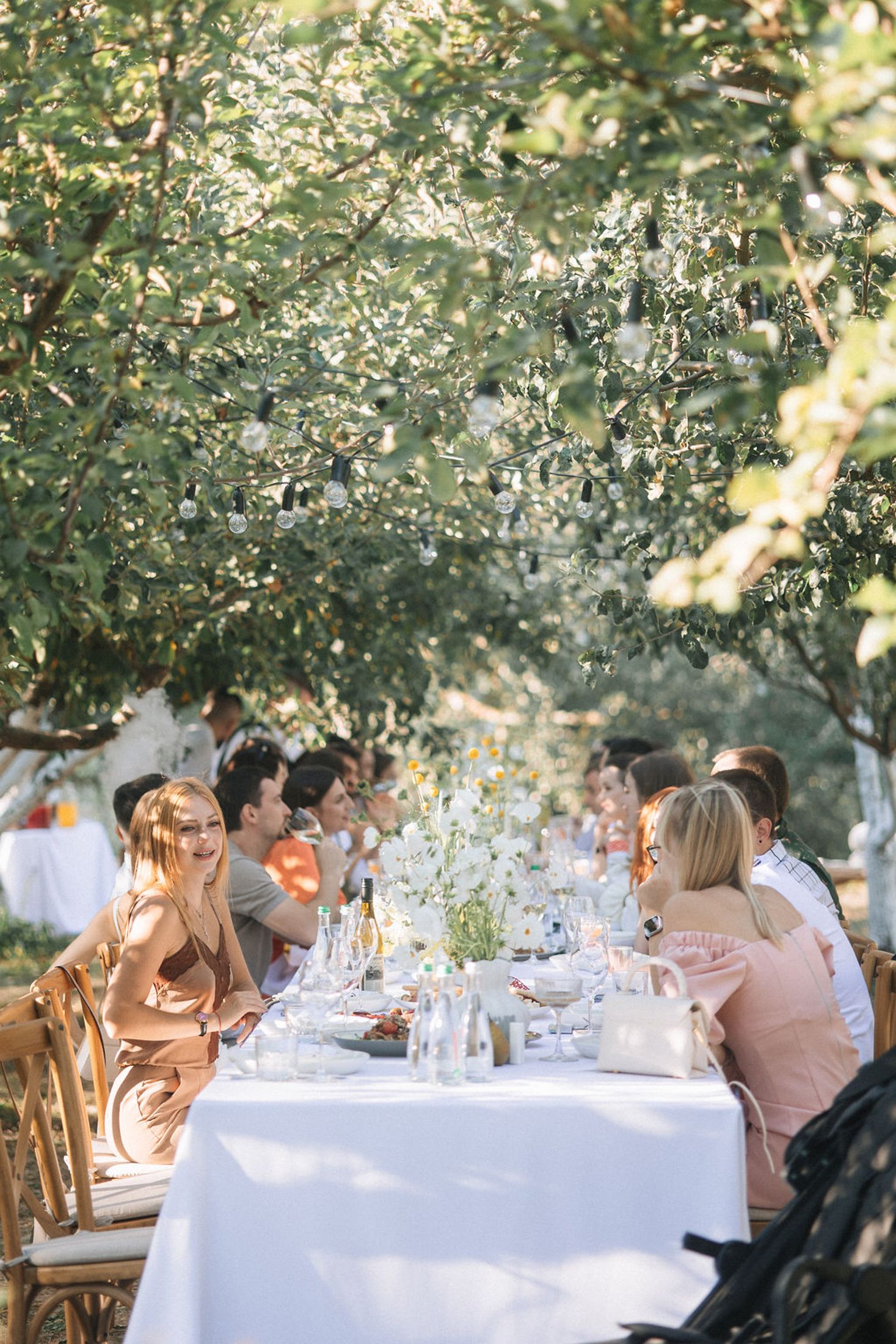 Guests seated at elegant white-clothed dining tables under trees at an outdoor garden party reception.