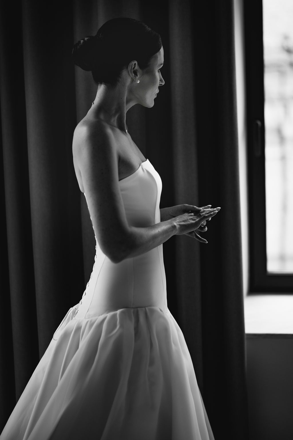 Black and white photo of someone in a white wedding dress checking their phone near a window.