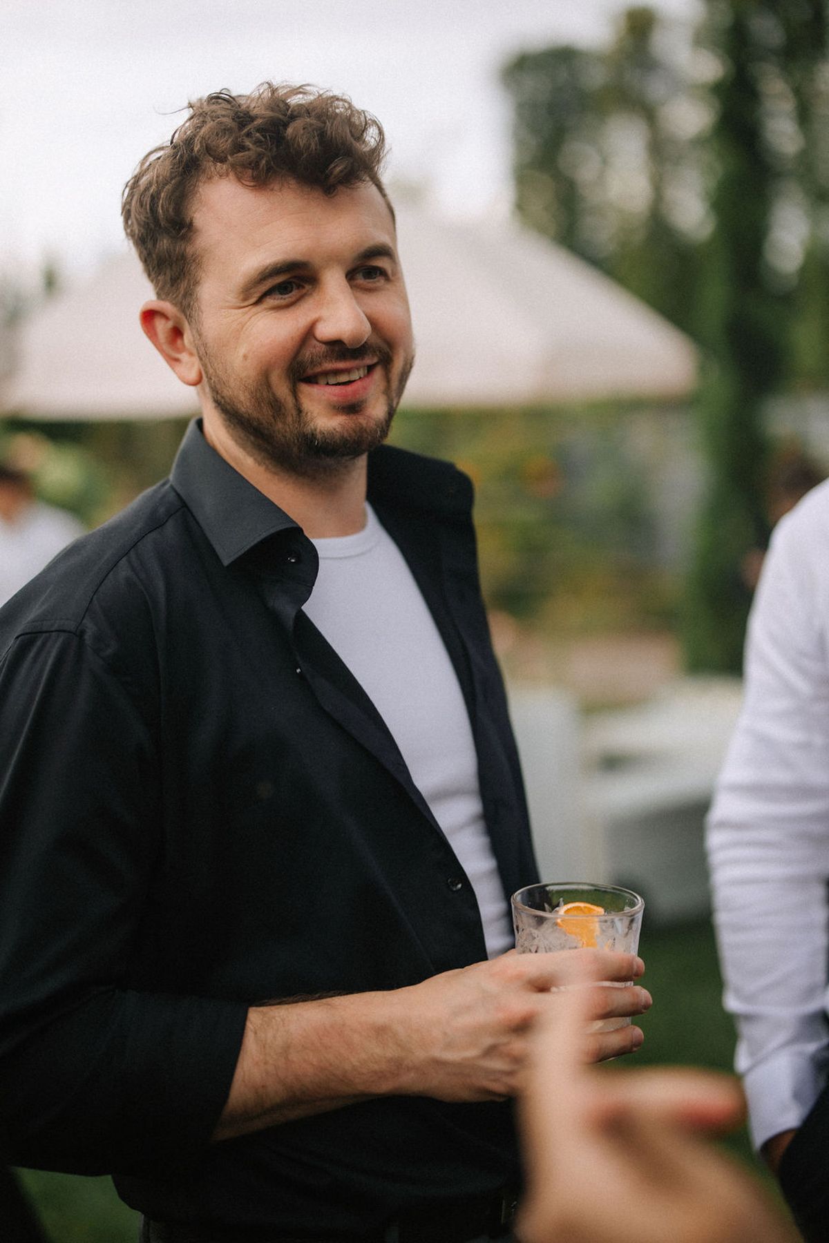 A person in a black jacket holds a drink while engaging in conversation at an outdoor gathering.