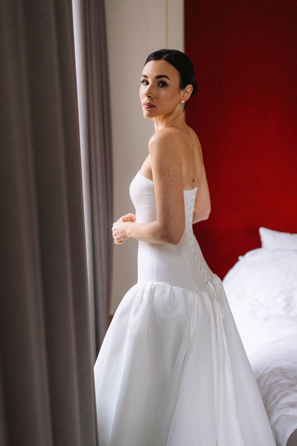 A bride in a classic white strapless wedding gown stands by a window with red walls and gray curtains in the background.