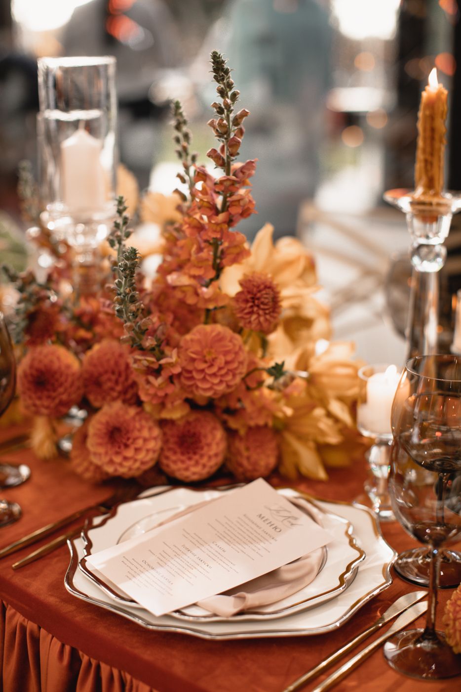Autumn-themed place setting with coral flowers, tall candles, and elegant dinnerware on rust-colored tablecloth.