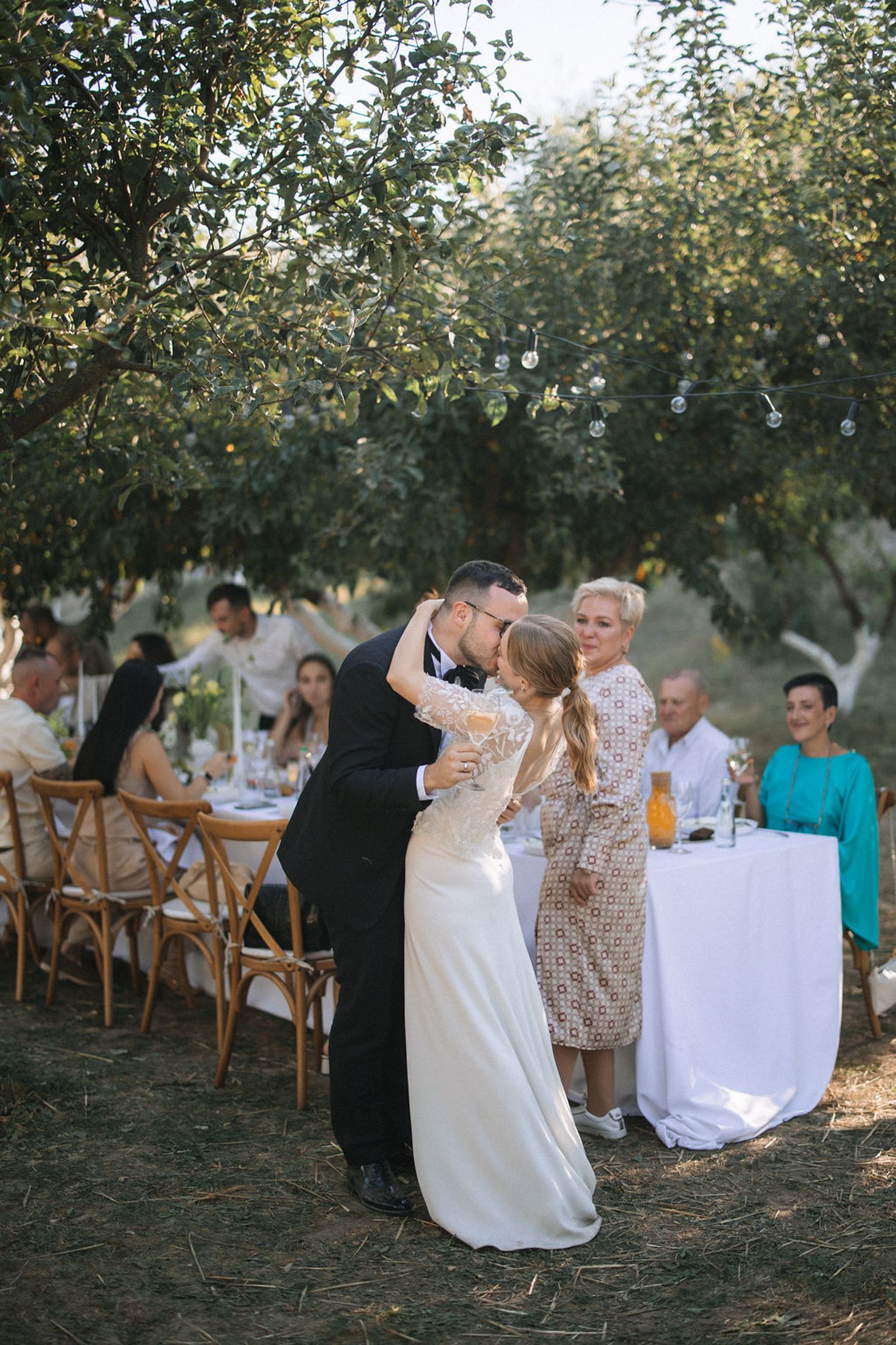 Guests enjoying an elegant outdoor dinner party under trees at sunset with rustic wooden chairs and white tablecloths.